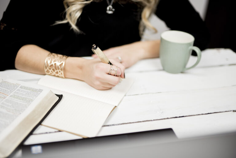 A woman writes in a lined notebook beside an open book and green mug on a white wooden desk