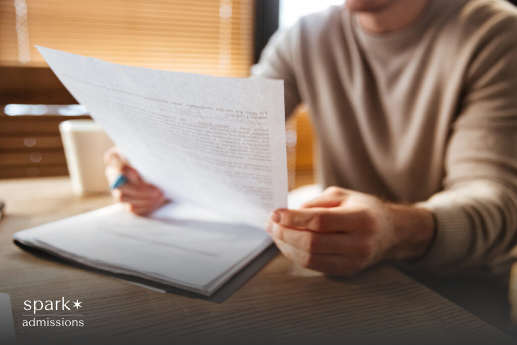A student sits at a desk holding a printed essay or document with a notebook in front