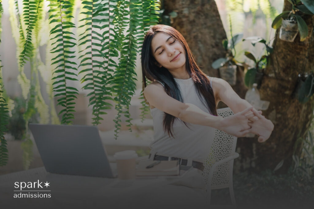 A woman stretches her arms at an outdoor table while working on a laptop surrounded by greenery