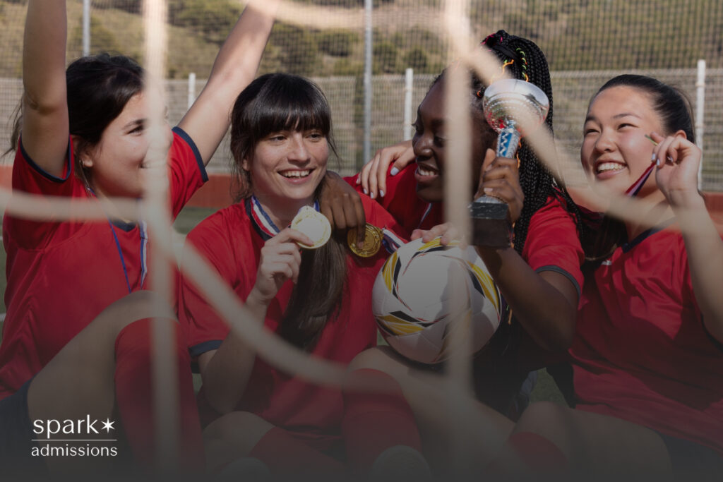 A group of girls in red soccer uniforms celebrate with medals, a trophy, and a ball on the field