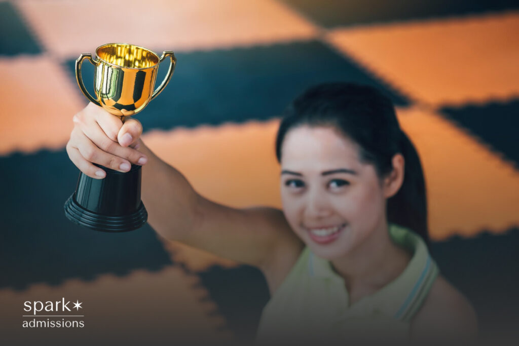 A smiling girl proudly raises a gold trophy toward the camera on a gym floor