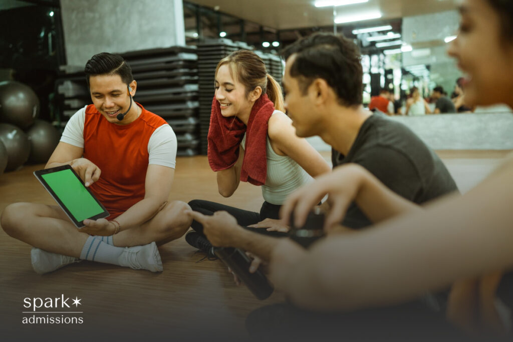 A fitness instructor shows a green-screen tablet to a group of smiling young adults sitting on a gym floor