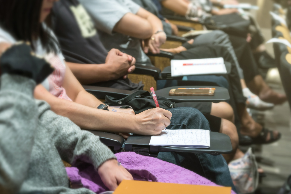 Students sit in a lecture hall taking notes on notebooks with pens during class