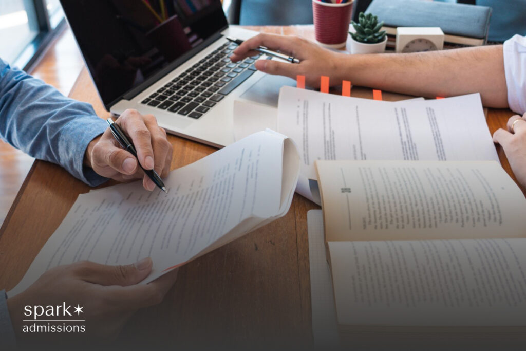 Two people review printed documents and take notes next to a laptop on a wooden desk