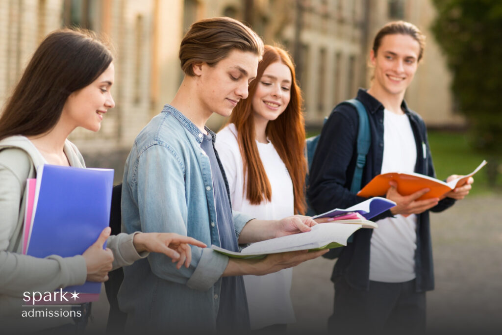 Four students walk on campus while talking and looking at open books and folders