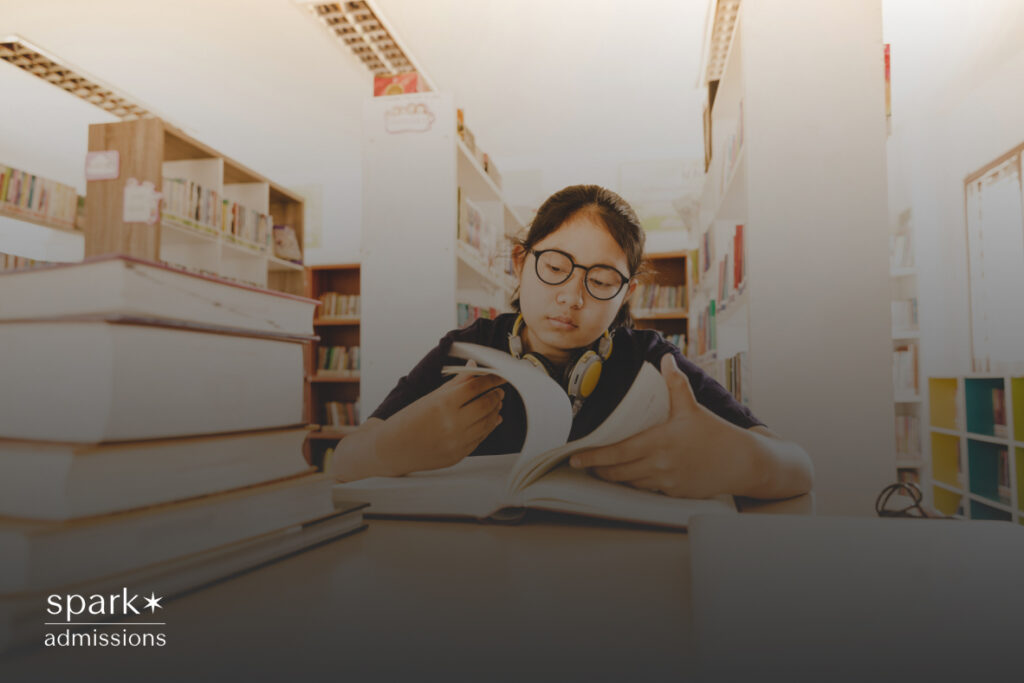 A student wearing glasses and headphones reads a book at a table surrounded by library shelves