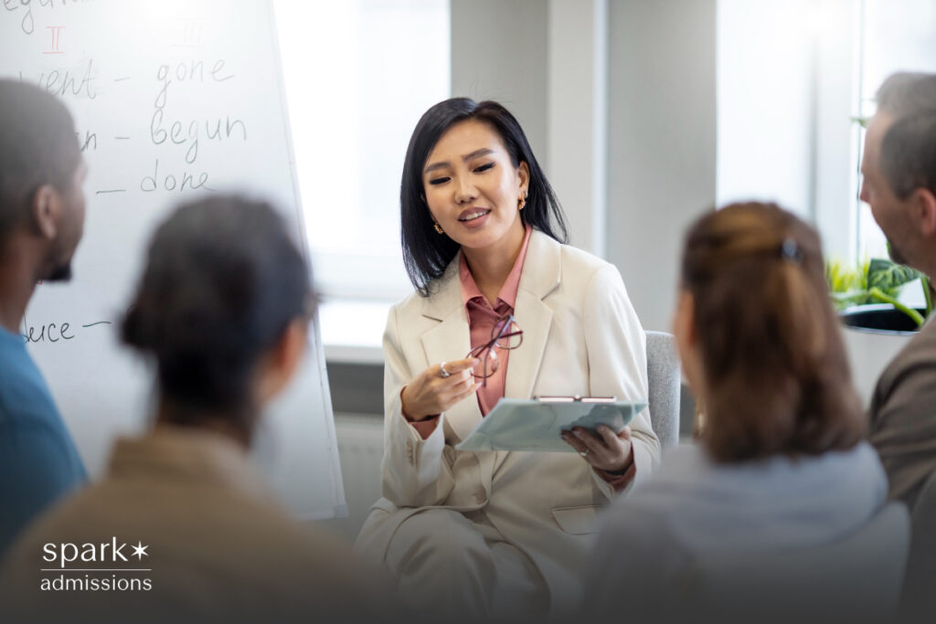  An Asian teacher in a cream blazer leads a discussion with adult learners in a classroom with a whiteboard