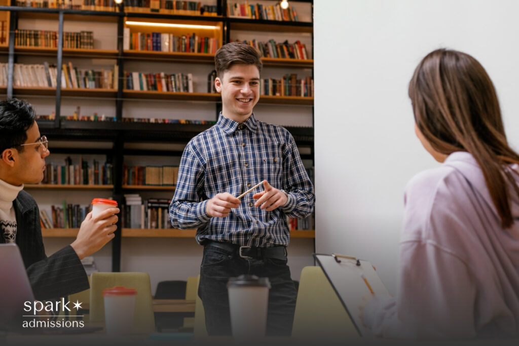 A smiling student stands in front of a bookshelf giving a presentation to classmates
