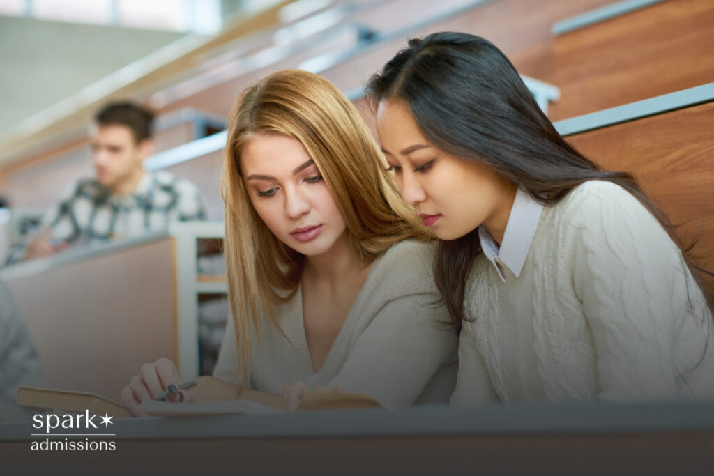 Two female students sit in a lecture hall, reading from a book and focusing on study materials