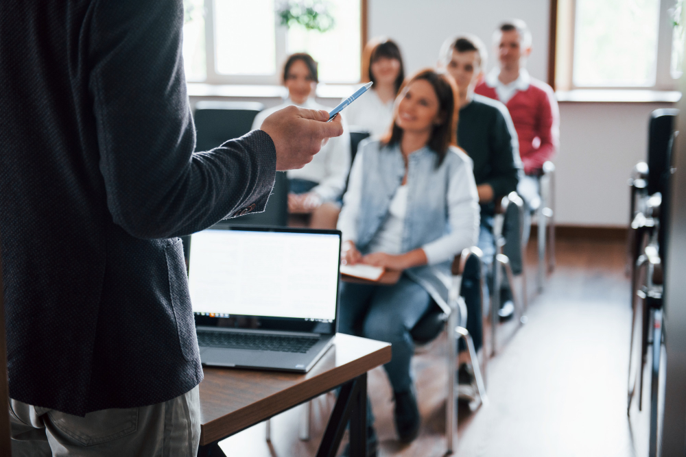A presenter stands in front of a group of students seated in rows, holding a pen beside a laptop