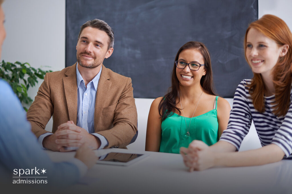 A group of three young adults sit across a table, smiling and engaged in a conversation during a college admissions meeting
