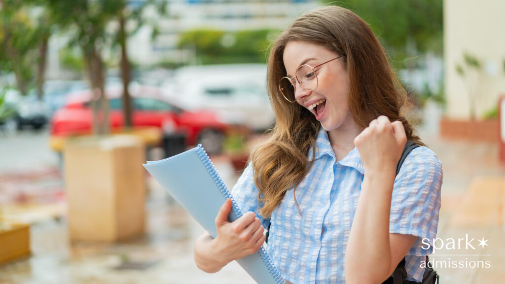 A smiling student pumps her fist while looking at a blue spiral notebook outdoors