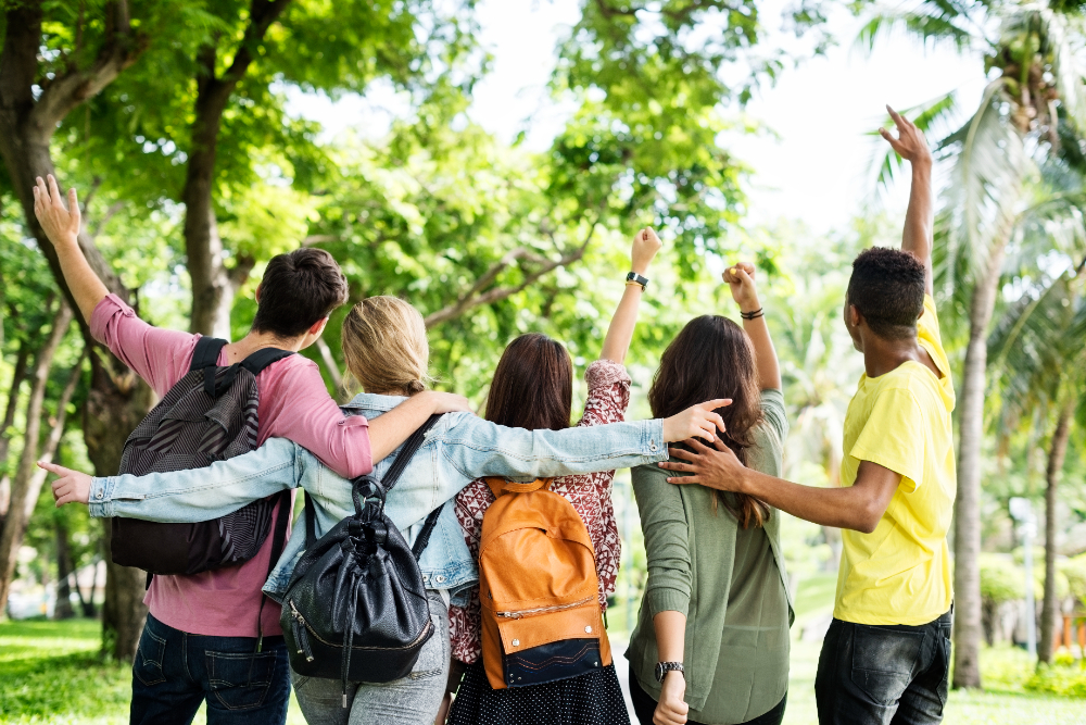 Group of five students with backpacks walking through a park with arms raised, seen from behind