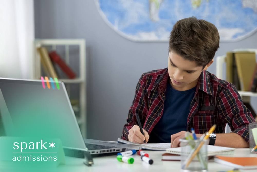 High school student studying at a desk with a laptop, writing notes and completing homework in a home study space.