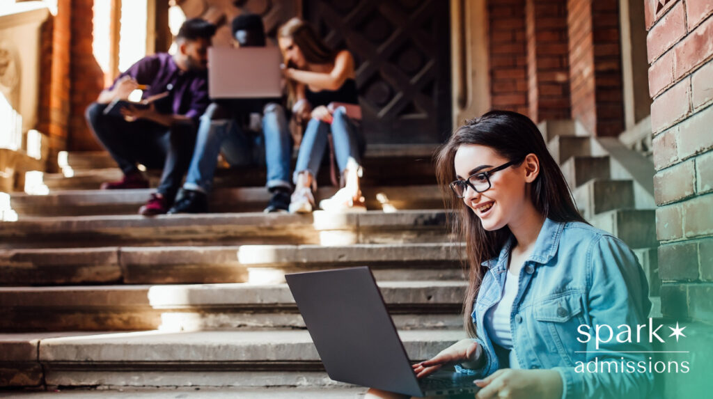 A smiling student using a laptop on campus steps with a group of students talking in the background