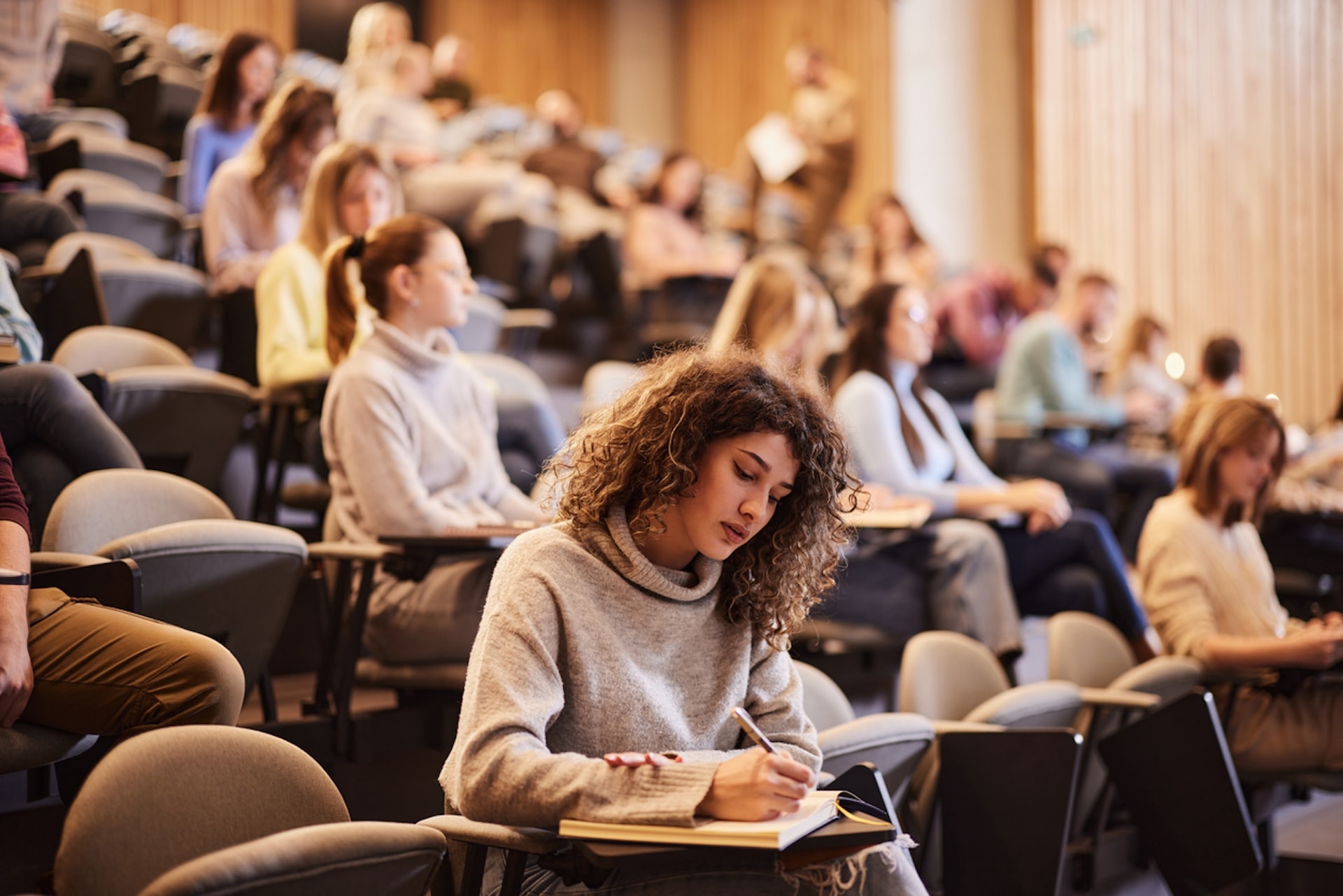 Female college student writing an exam during a class at lecture hall. Her classmate are in the background