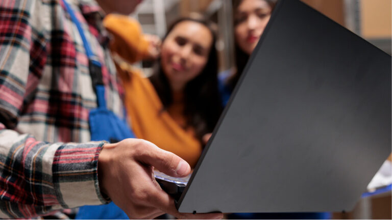 A student holds a laptop while others in the background observe and listen in a casual group setting
