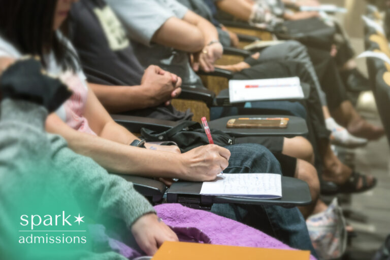 College students taking notes during a classroom lecture while seated in a university lecture hall.