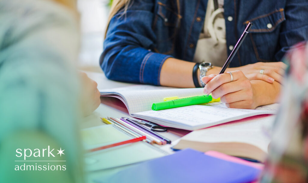 A student writes in a notebook using a pen with a green highlighter and open books nearby