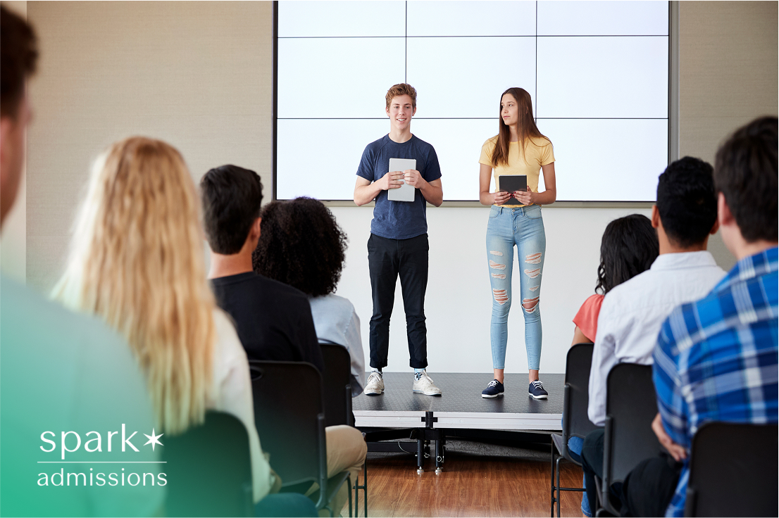 Two students giving a presentation on stage in front of classmates in a school auditorium