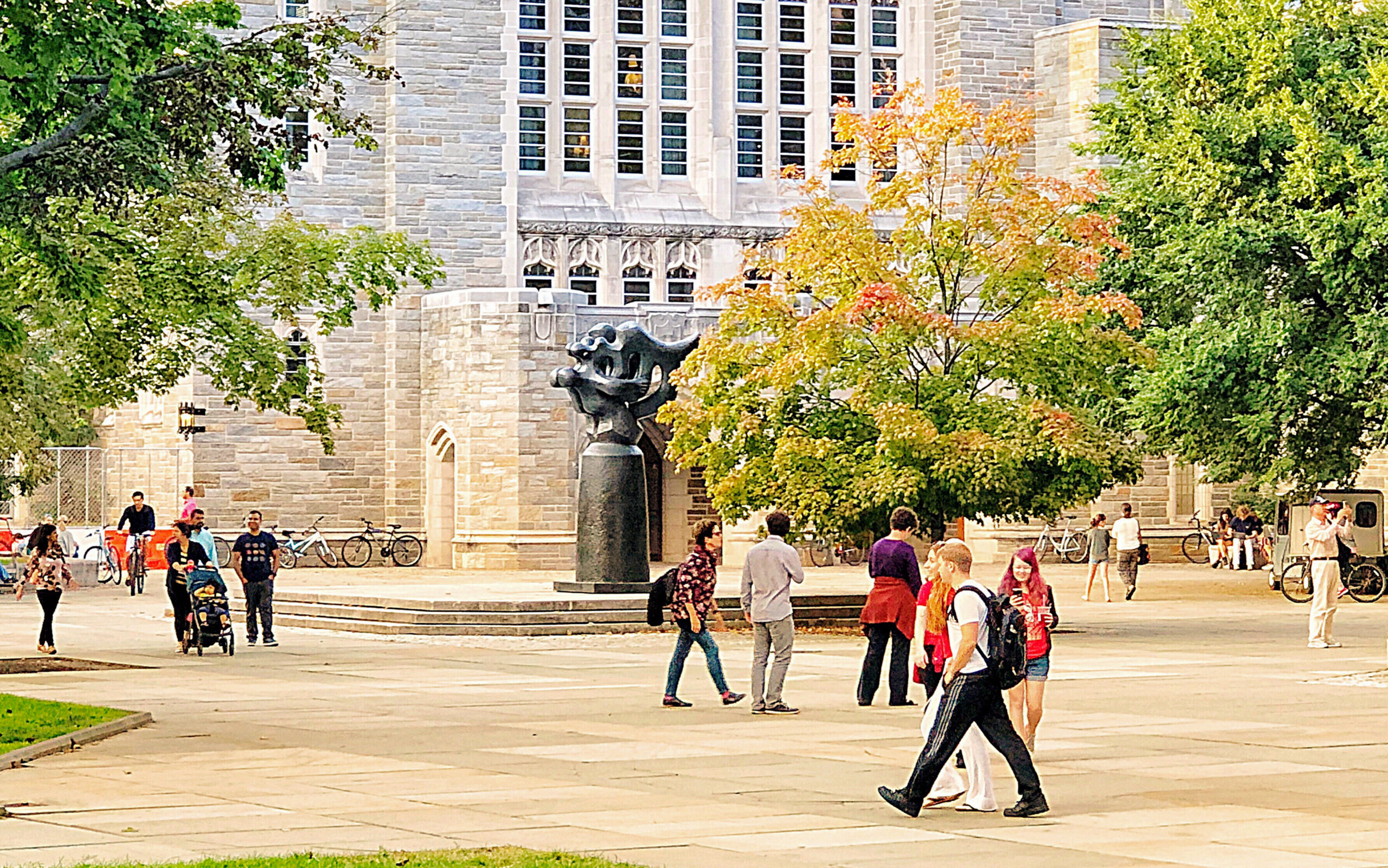 College students walking across a university campus with bikes, trees, and historic buildings in the background