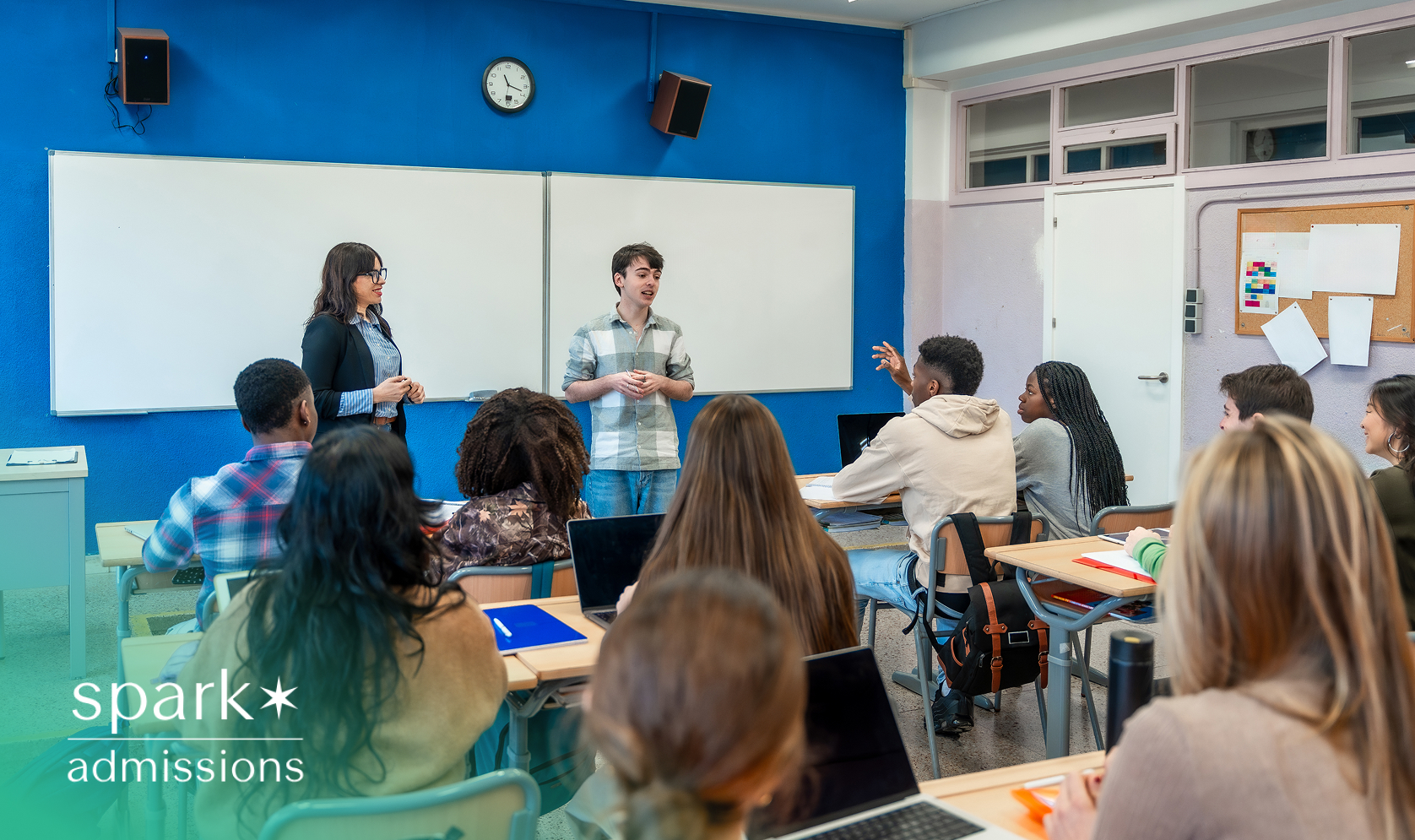 Teacher and student standing at the front of a classroom while students listen and engage in discussion