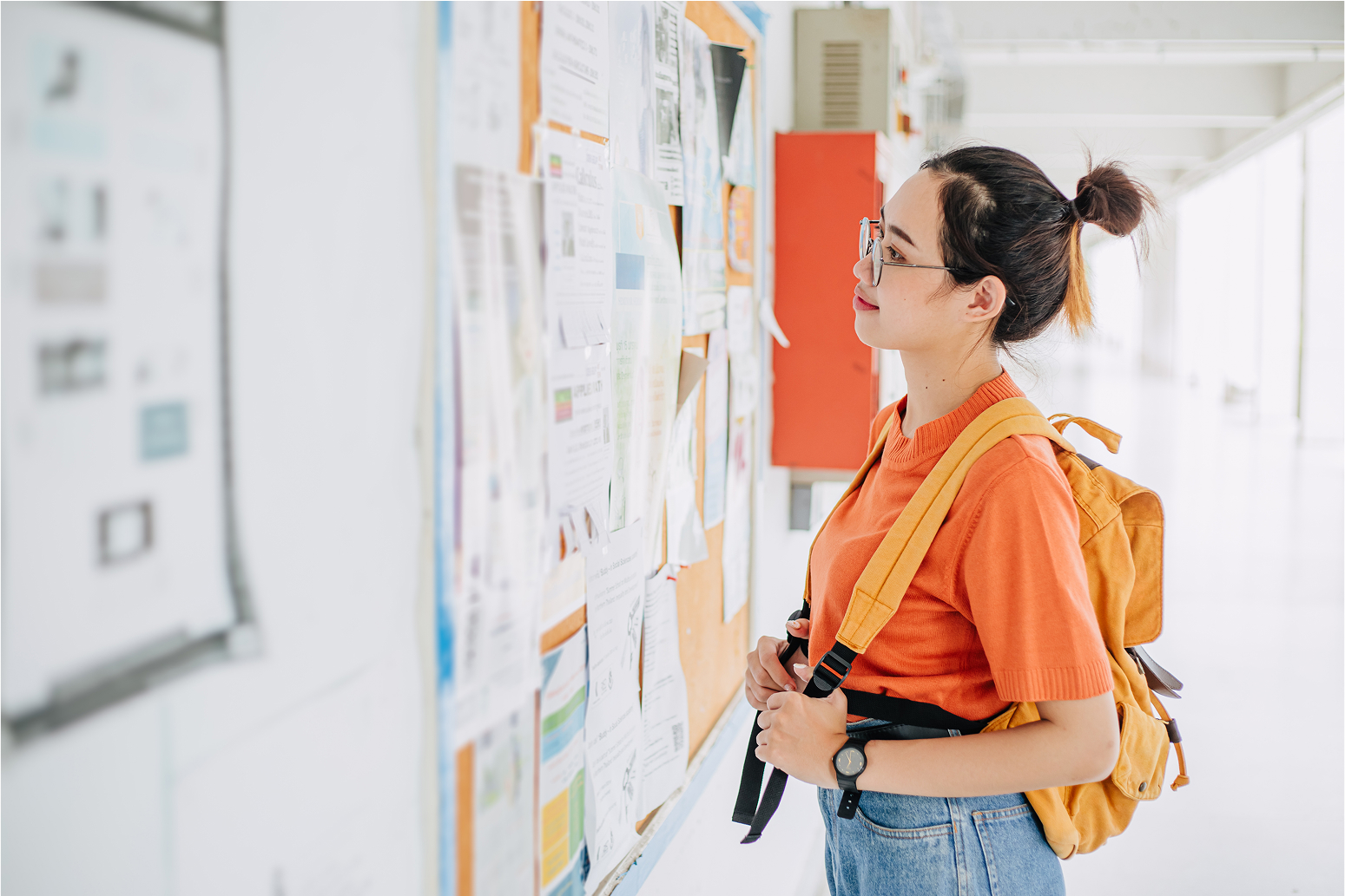 A student in orange top with a yellow backpack reading a bulletin board in a school hallway