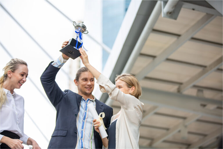 A man holding up a trophy as two women celebrate beside him outside a modern building
