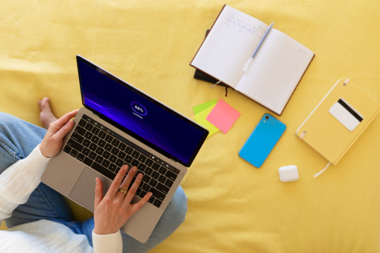 A person using a laptop surrounded by notebooks, phone, and sticky notes on a yellow bedspread