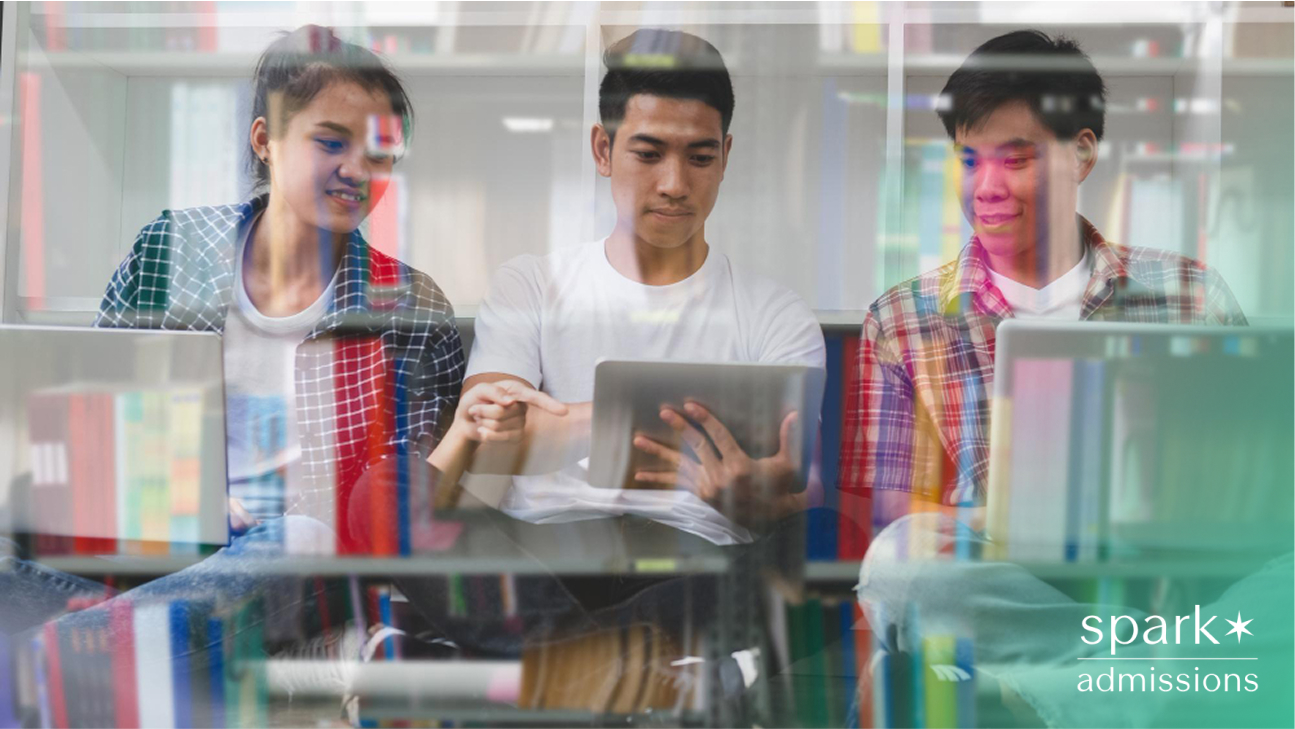 Three students sitting together using laptops and a tablet, viewed through a glass bookshelf