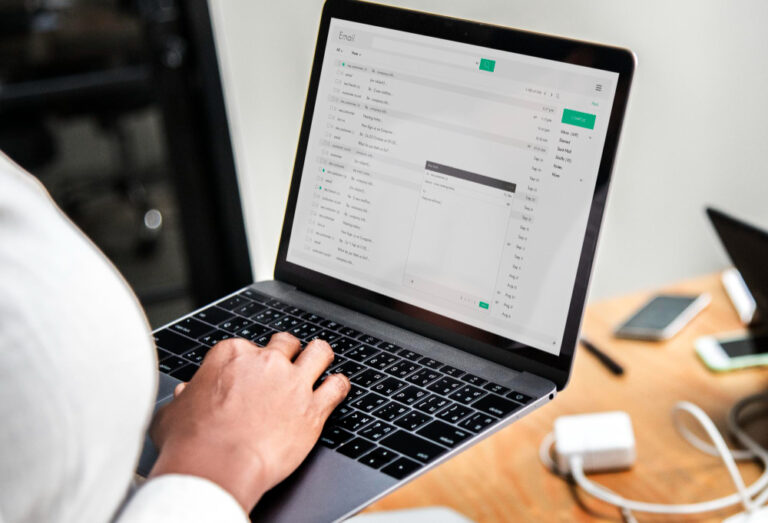 A person checking and typing emails on a laptop at a desk with phone and charger nearby