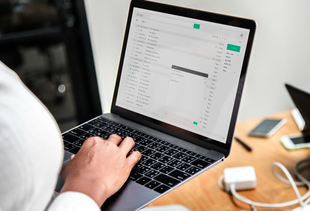 A person checking and typing emails on a laptop at a desk with phone and charger nearby