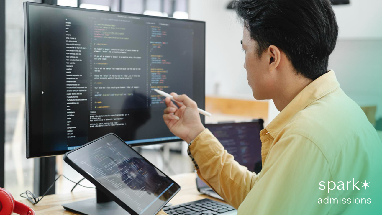 A student pointing at code on a monitor while working on multiple screens in a computer lab