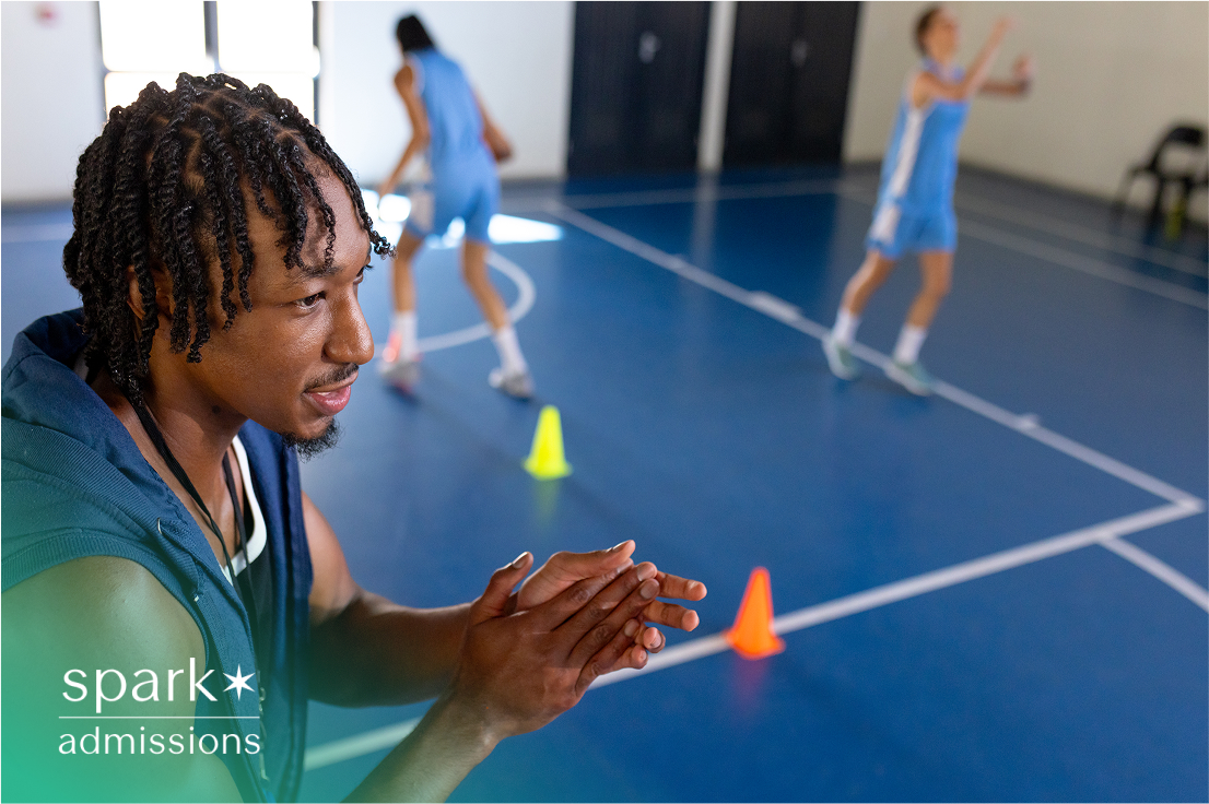 A basketball coach clapping on the sideline as players run drills in the gym