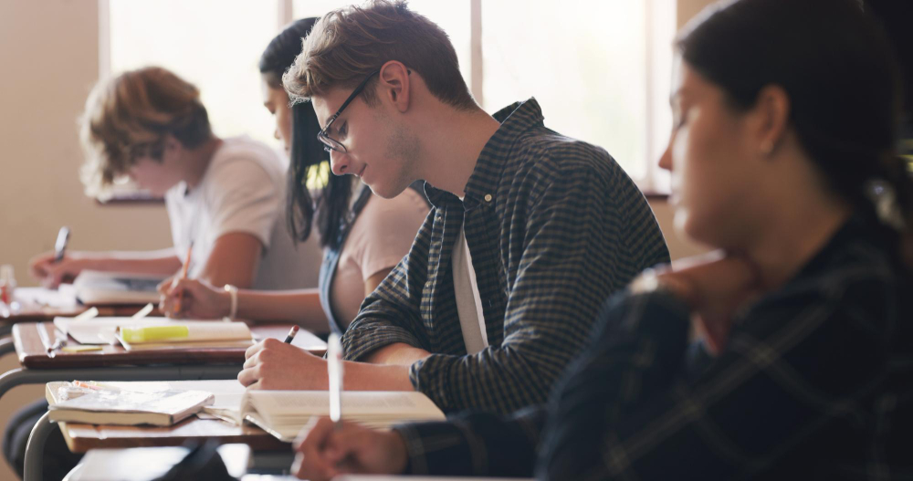 Focused student sitting at a desk, writing answers on an exam sheet in a classroom setting.