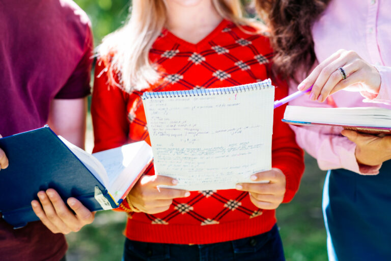 Three students holding notebooks and textbooks, reviewing handwritten notes together outdoors