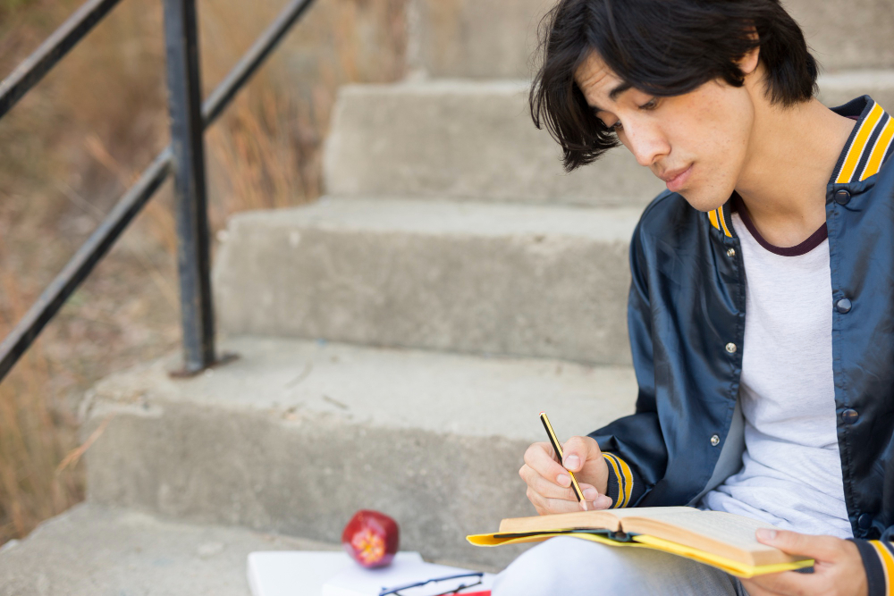A student sitting on outdoor steps, writing in a notebook with books and an apple beside him