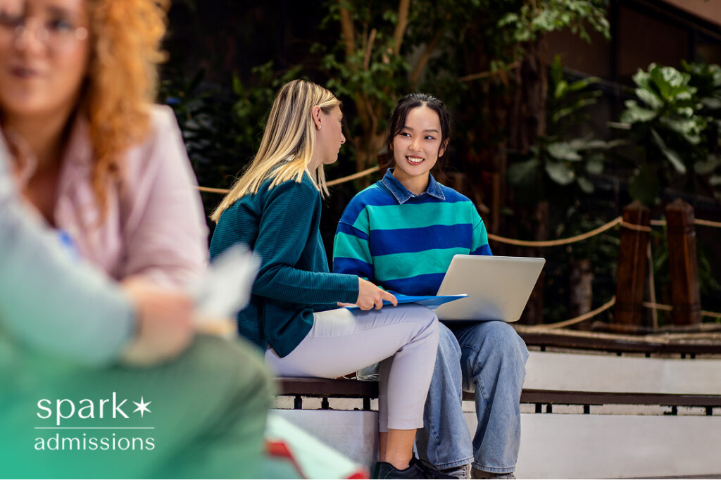 Two college students sitting outdoors on campus, smiling and working together on a laptop, representing collaborative learning and campus life
