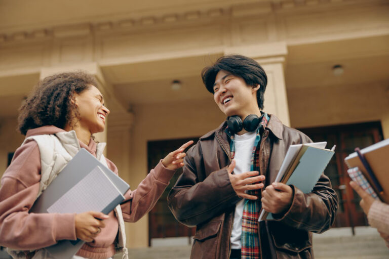 Two college students smiling and talking while holding notebooks outside a campus building