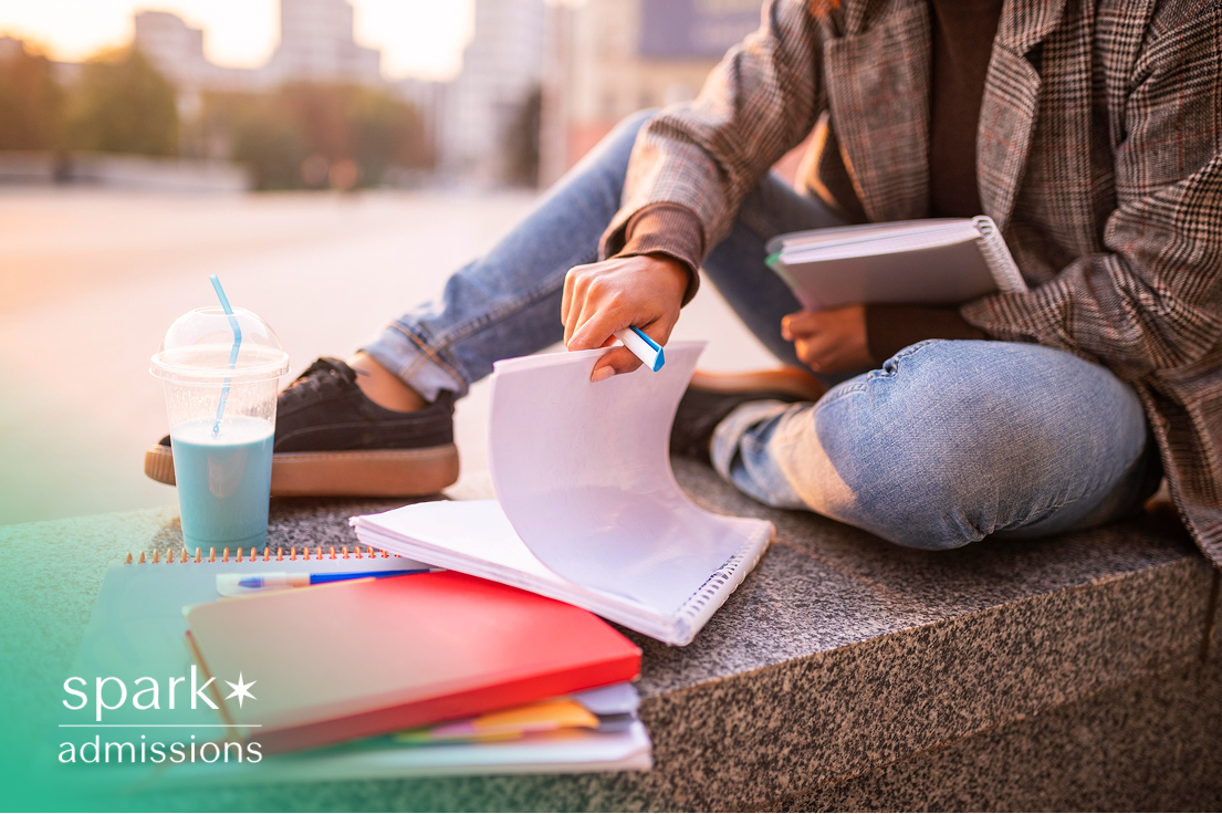 A student sitting on a concrete bench outdoors, flipping through notebooks with a drink nearby