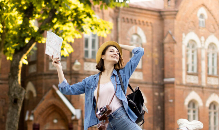 Smiling student holding a map, wearing a hat and backpack in front of a historic college building