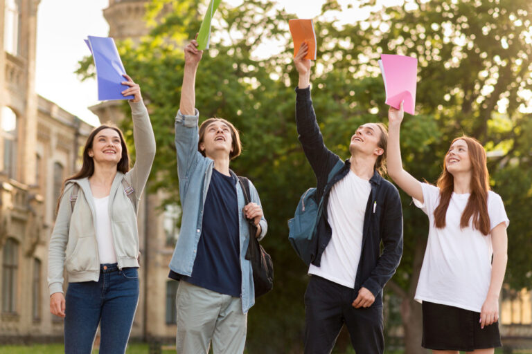 Four smiling students outside holding colorful folders in the air in front of a college building