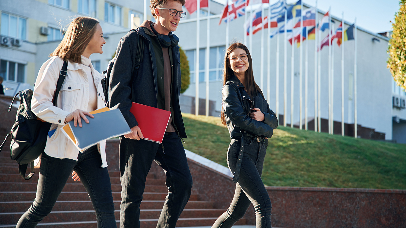 Three students walking down steps outside a university with international flags in the background