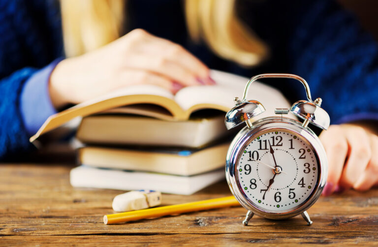 Alarm clock on a wooden table in front of stacked books and a person studying