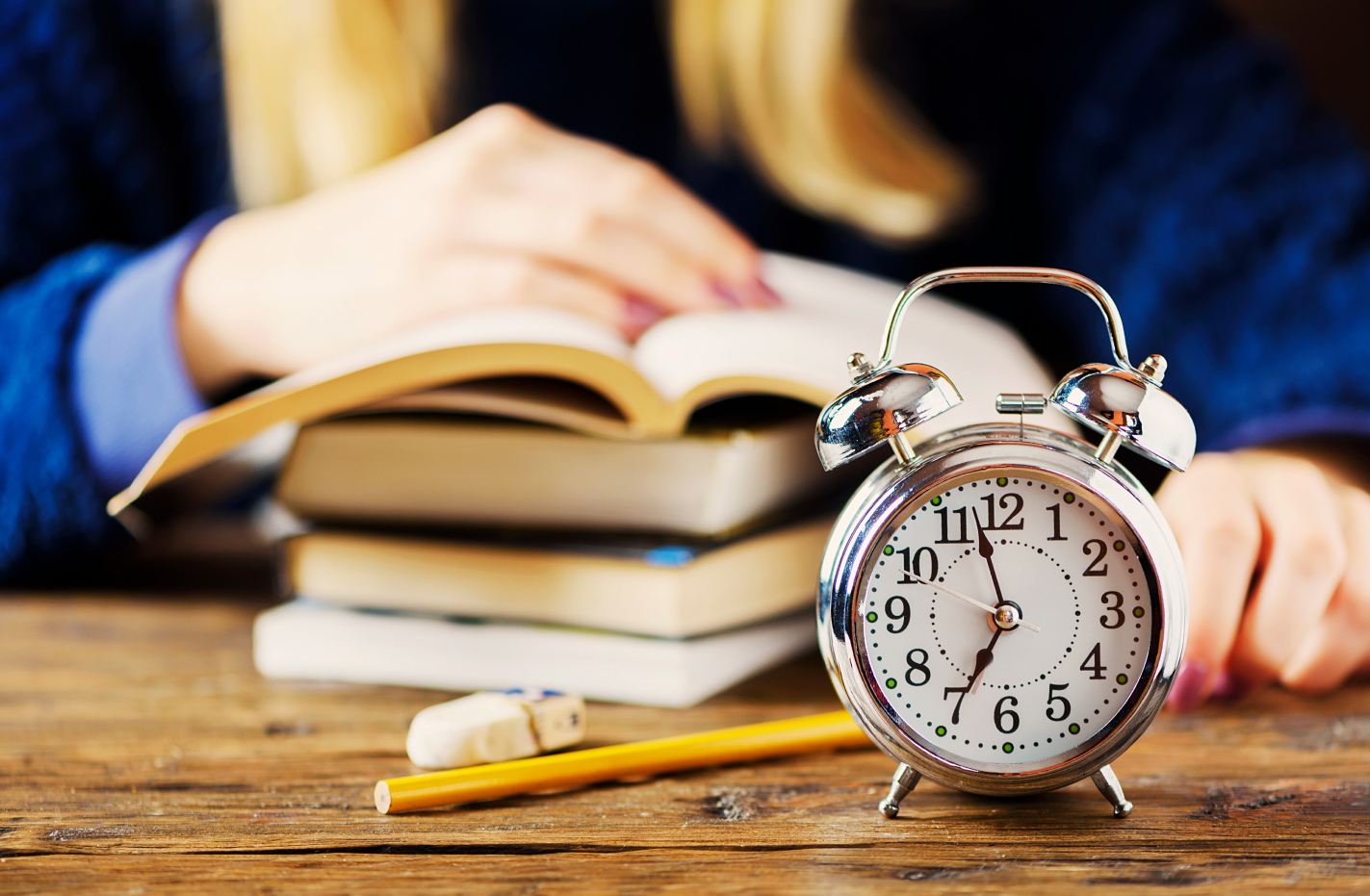 Alarm clock on a wooden table in front of stacked books and a person studying