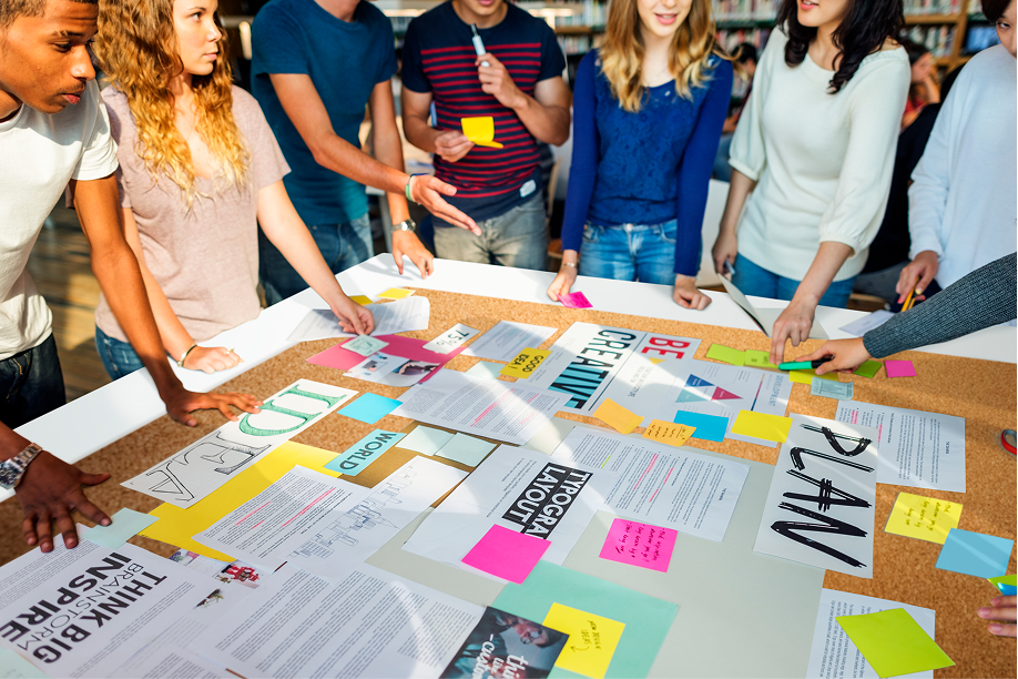 Group of students gathered around a large table filled with notes, charts, and colorful post-its, actively organizing ideas for a project