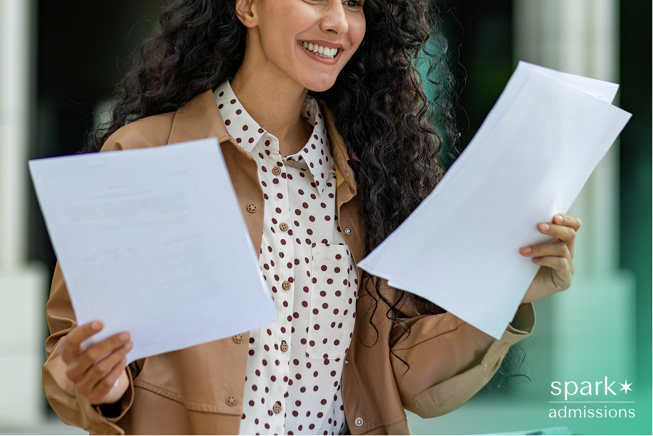 Smiling female student holding paperwork, wearing a polka dot shirt and tan jacket