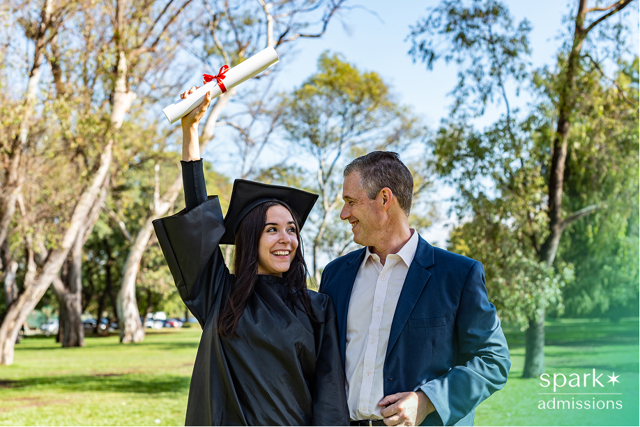 Young woman in graduation gown holding diploma beside a man in a suit outdoors