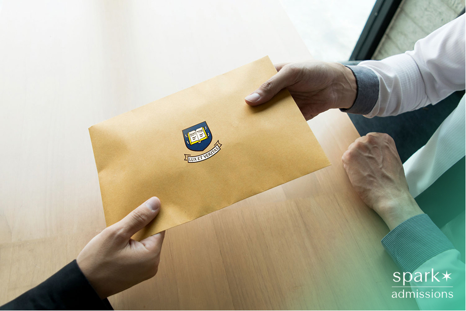 Two people exchange a large envelope with the Yale University logo on a wooden table