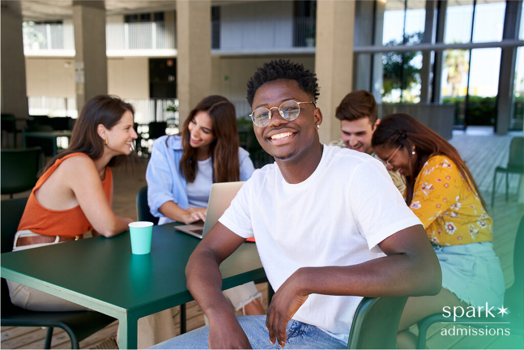 A smiling student sits at a table with friends working on laptops in a modern campus setting